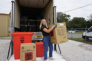 A worker smiling while handling biohazard boxes for medical waste management, loading them onto a truck with safety containers.